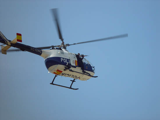 Helicóptero de Policía Nacional en vuelo, y un Policía Nacional con un arma en el exterior del mismo, prestando apoyo aéreo.