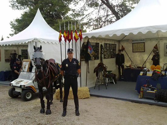 Un policía nacional posa junto a una maqueta de un caballo y una caseta donde se exhibe diverso material de unidad especial de caballería.