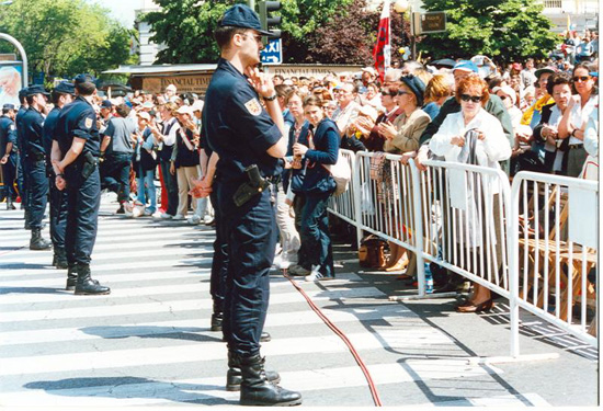 Formación en línea de varios policías protegiendo un desfile. Delante de ellos hilera de vallas protectoras tras las que se encuentran los asistentes.