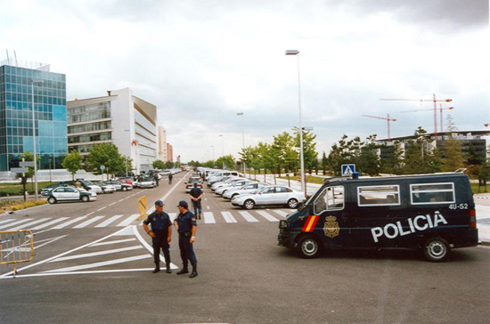 Tres policías junto con un furgón de la Unidad cortando el acceso a una calle en la que se observan vehículos de Guardia Civil y particulares.