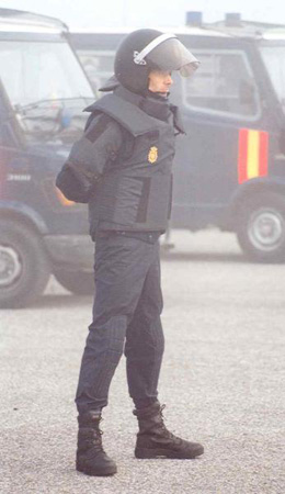Policía con chaleco y casco puesto, con los brazos a la espalda, frente a dos furgones. Se observa ambiente denso por humo.