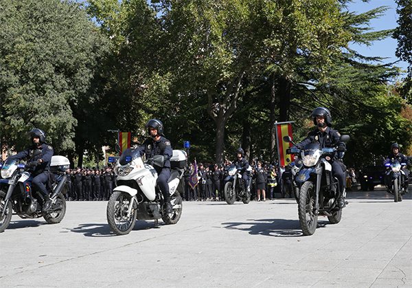 Desfile motorizado de vehículos policiales. Al frente 6 motocicletas. Le siguen otros vehículos. De fondo policías en formación.