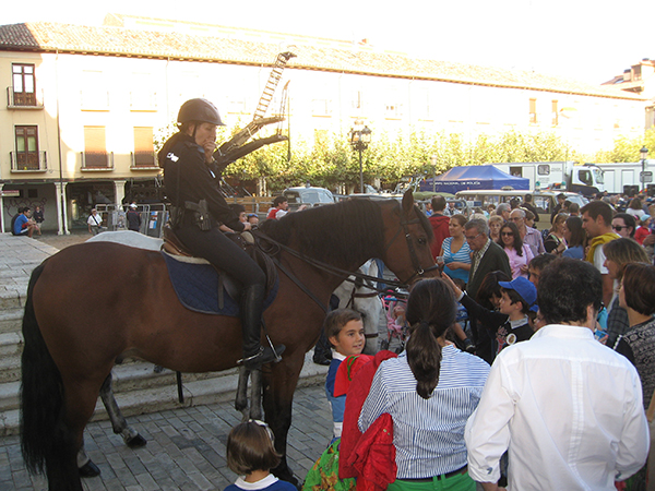 Amazona uniformada sobre su caballo color marrón. Se encuentra rodeada de gente. Un niño acaricia el morro del caballo.