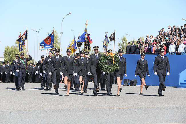 Desfile de uniformados portando distintas banderolas de unidades policiales. Al frente hombre y mujer portando corona de flores con bandera de España.