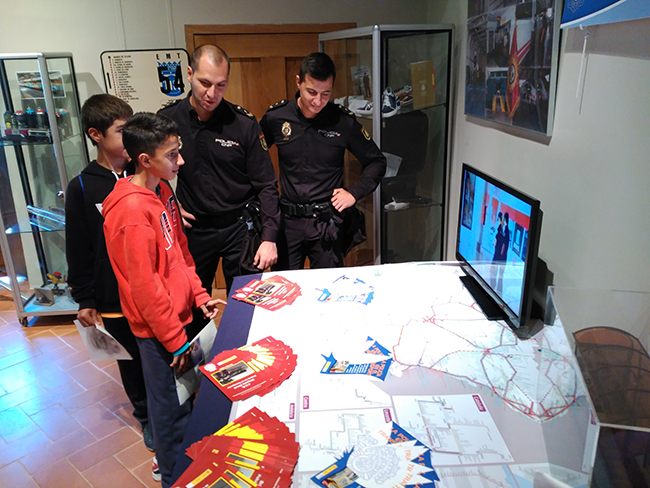 Dos niños atienden a la explicación de dos policías sobre lo que se ve en un monitor. Alrededor de éste se encuentran varios folletos informativos.