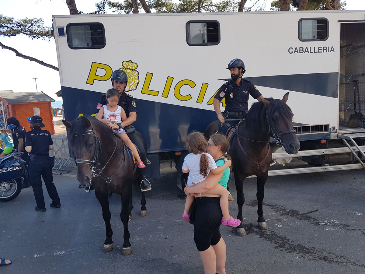 Dos policías de la Unidad de caballería, uno de ellos con una niña montada con él y una señora,  con niña en brazos, observando la escena