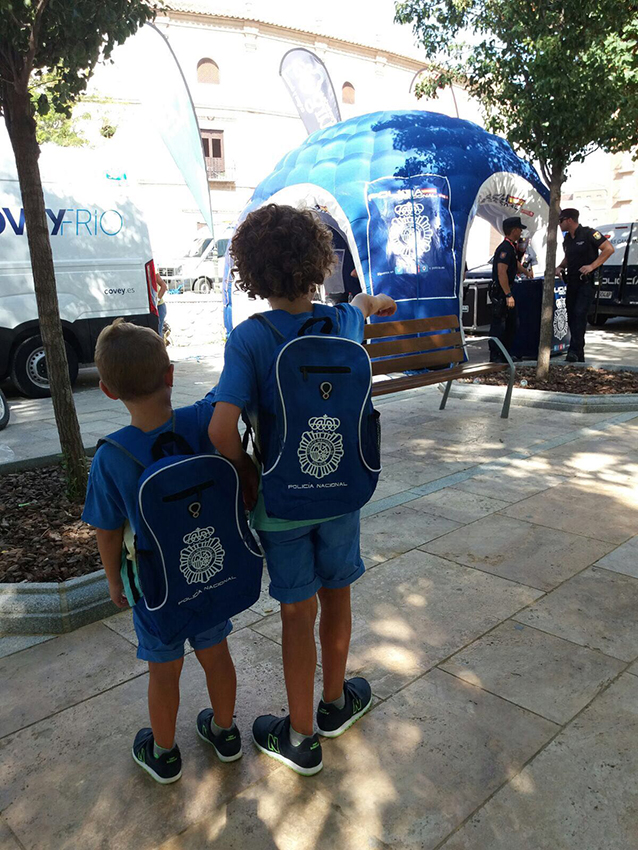 En el frente dos niños con dos mochilas con logo de la policía y al fondo carpa de la Policía Nacional
