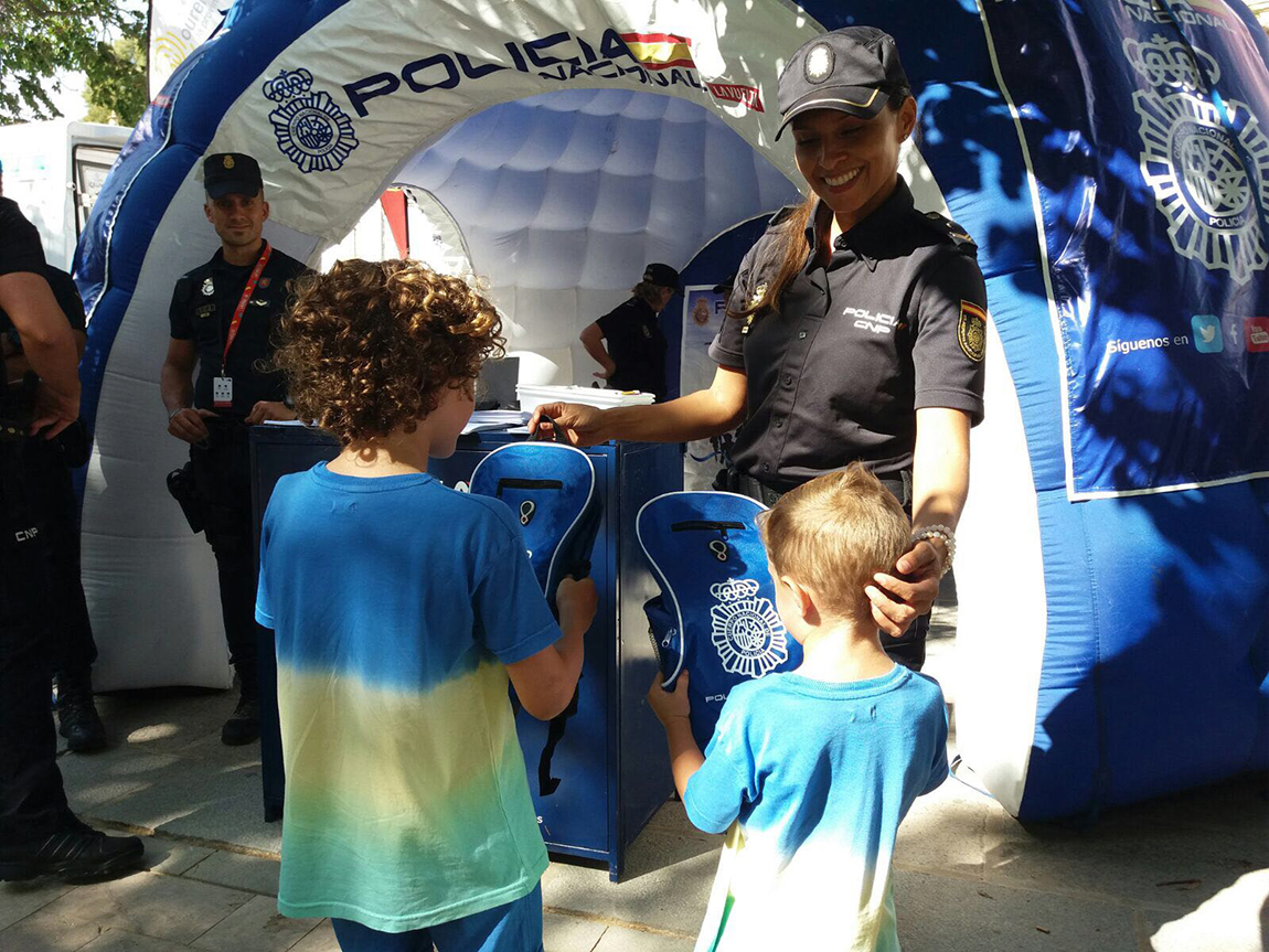 En el centro una agente de policía entregando dos mochilas con el logo de la policía ante la carpa informativa