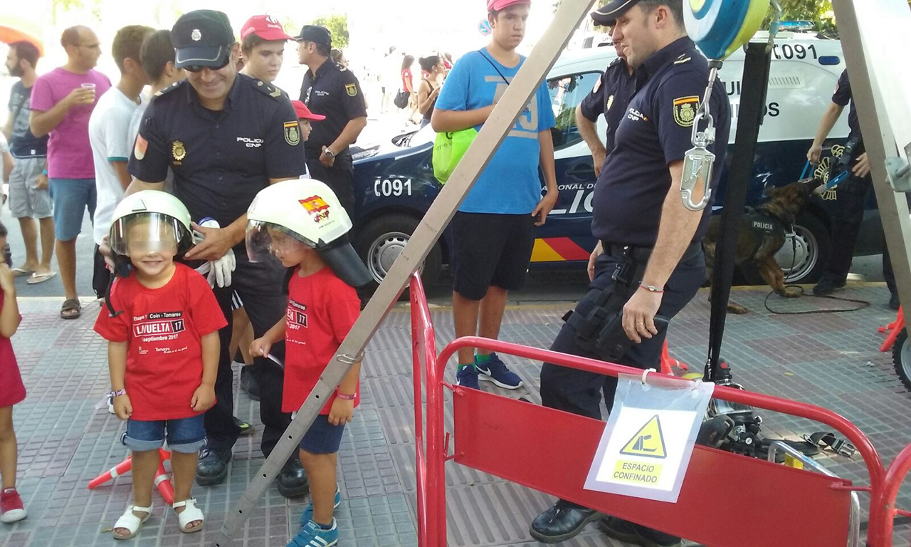Un policía colocando un casco de trabajo a un niño, junto a ellos otro pequeño con casco y un agente mirando a los niños