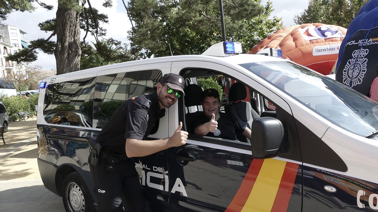 Un agente con un joven que se encuentra dentro de vehículo policial saludando y con camiseta con el logo de la policía