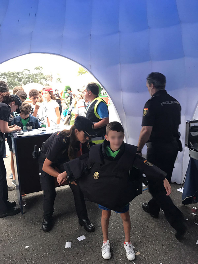 Una agente de policía, dentro de la carpa informativa,  ayudando a un niño a ponerse un chaleco reglamentario 
