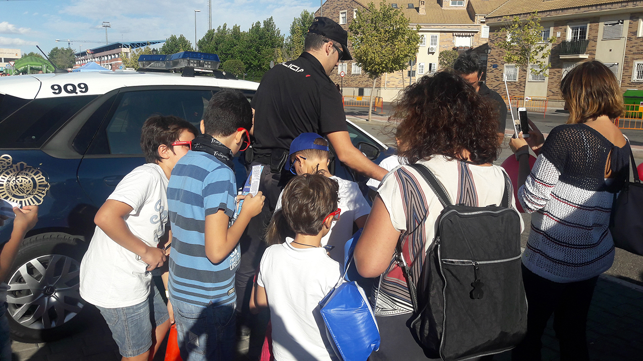 Varios niños y dos adultos rodeando a un agente de policía junto a un vehículo policial
