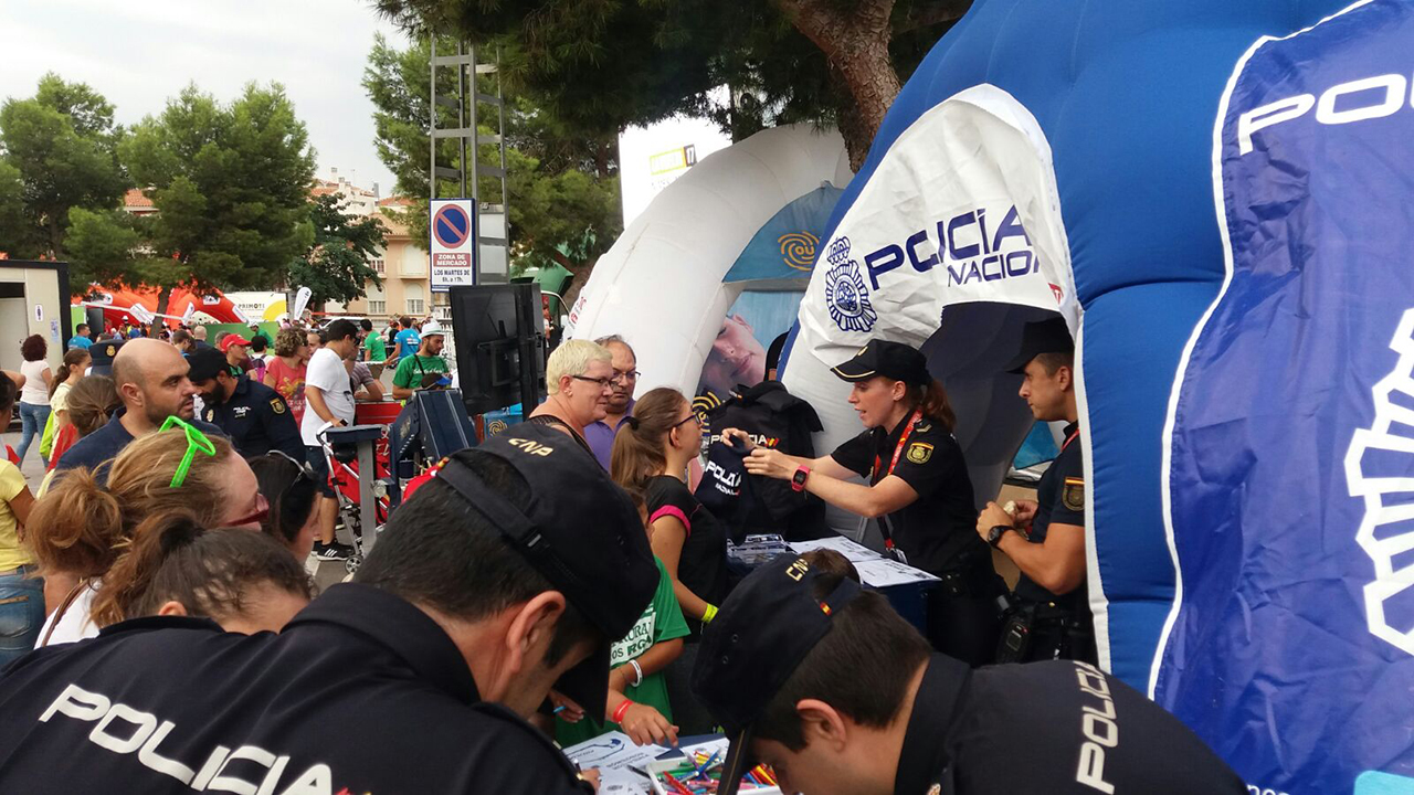 Carpa de la policía rodeada de visitantes y agente entregando camiseta de la policía a una niña, al frente otros agentes atendiendo a unos niños
