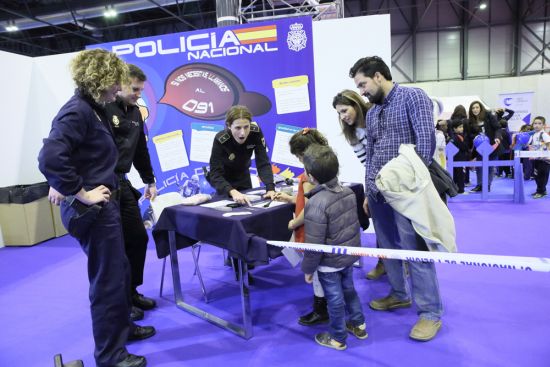 Dos niños recibiendo información, por parte de una agente de policía , observados por otros dos agentes y dos adultos.