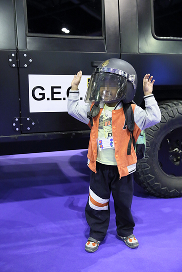Niño posando, con los brazos levantados y con casco del Grupo Especial de Operaciones (G.E.O.), detrás un vehículo de intervención de este grupo.