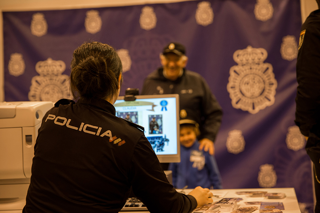 Agente de policía de espaldas, generando diploma acreditativo de asistencia a la feria, al fondo, niño con un adulto en el fotocol