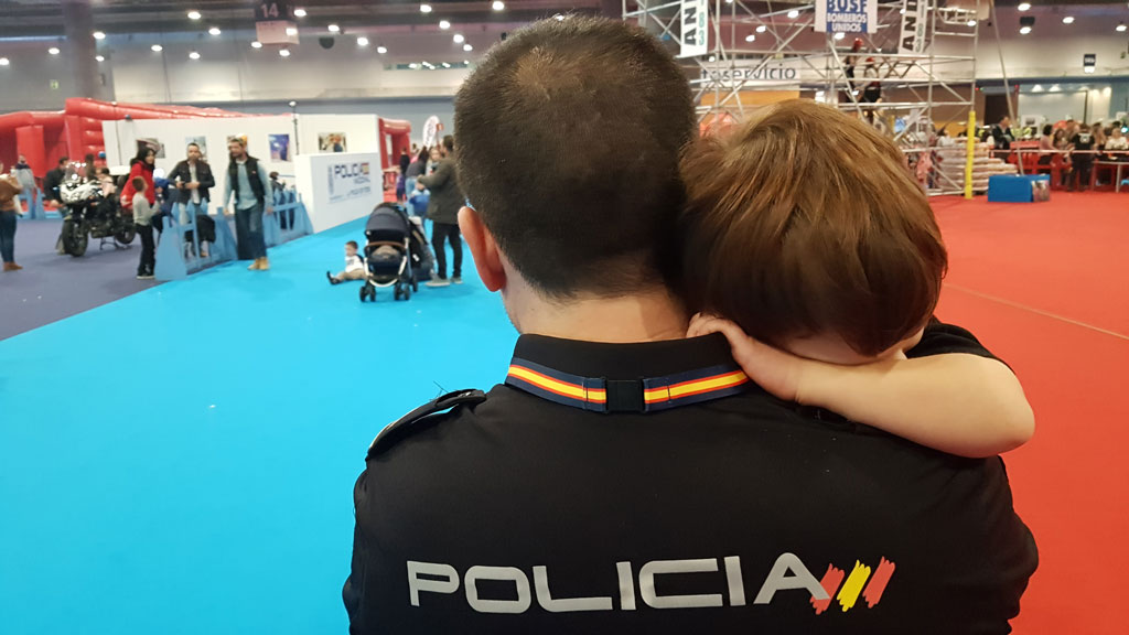 Primer plano, de espaldas, de un Policía Nacional portando a un niño entre sus brazos.
