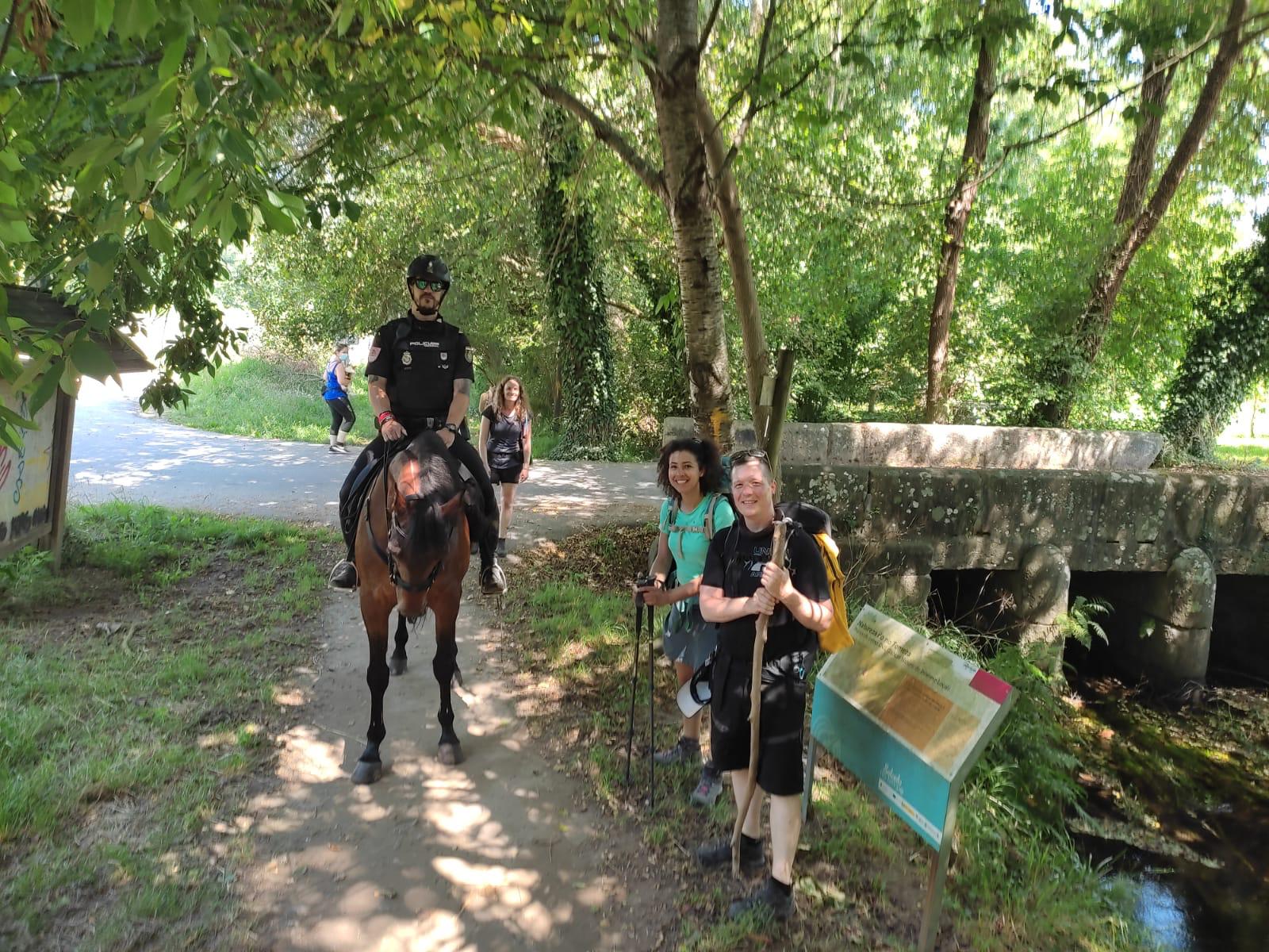 Un policía montado a caballo junto a dos peregrinos en un camino junto a un río y un puente.