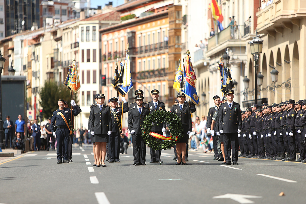 Formación de policías en posición de firmes. Encabezan la misma dos policías, hombre y mujer, portando corona con la bandera nacional.