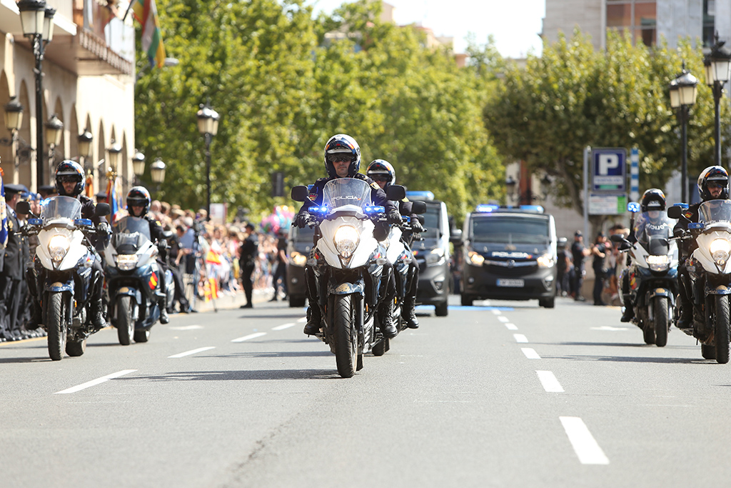 Seis agentes motorizados seguidos de furgonetas, en formación, en el desfile de la Policía Nacional. 