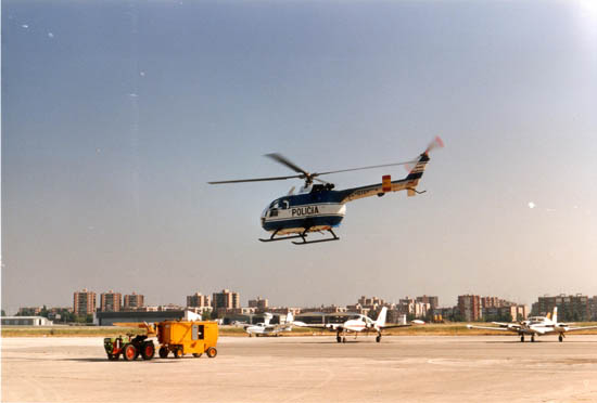Helicóptero de la Policía Nacional, modelo BO-105, realizando la maniobra de despegue, y de fondo varios aviones estacionados en la pista.