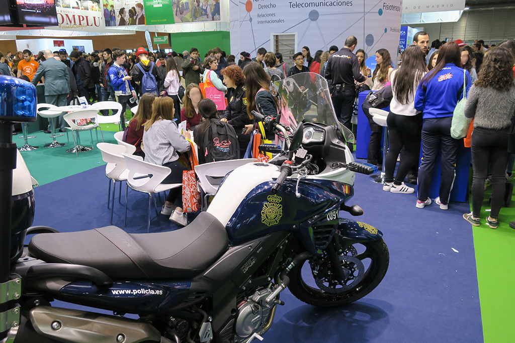 Motocicleta policial, detrás una mesa y mostradores,  con  jóvenes recibiendo información.