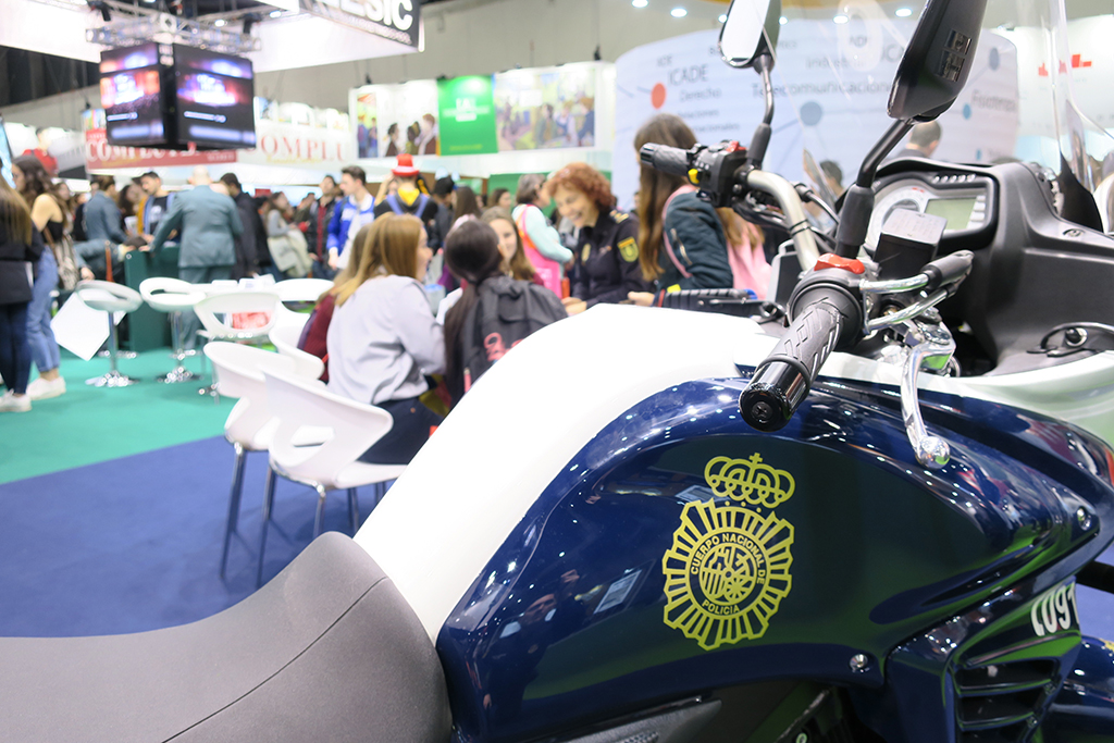 Primer plano del logo de la policía en la motocicleta, al fondo, jóvenes recibiendo información.