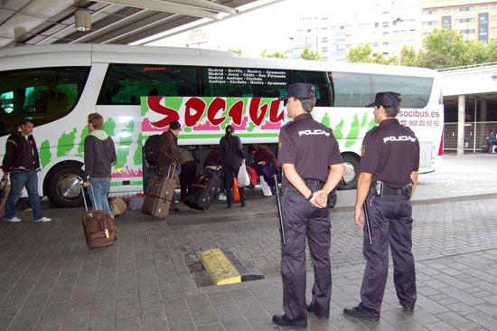 Dos Policías Nacionales vigilan el andén de una estación de autobuses, mientras viajeros introducen sus pertenencias en el maletero de un autobús.