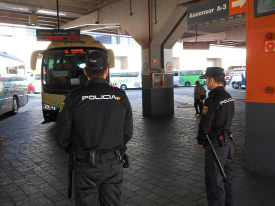 Dos Policías Nacionales vigilan el andén de una estación de autobuses, donde hay estacionados varios autobuses.