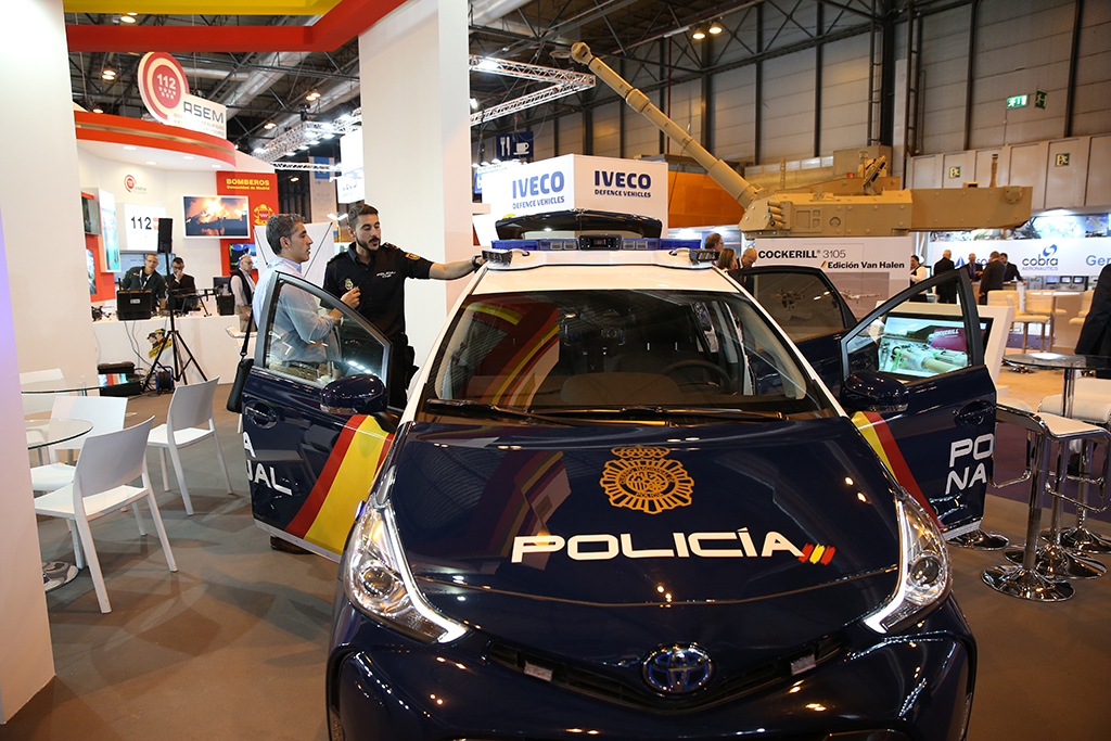 Policía mostrando a una persona el vehículo policial híbrido Toyota Prius, conocido como IZ. En el fondo, otros stands de la feria