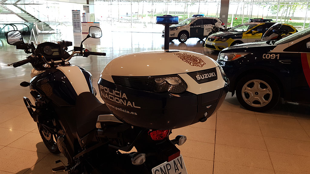 Motocicleta rotulada marca SUZUKI, con logo de la Policía Nacional y dirección web oficial de la policía. Se observan otros vehículos policiales.